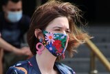 A woman wears a colourful mask, jacket, shirt and jeans as she walks past Flinders Street Station.