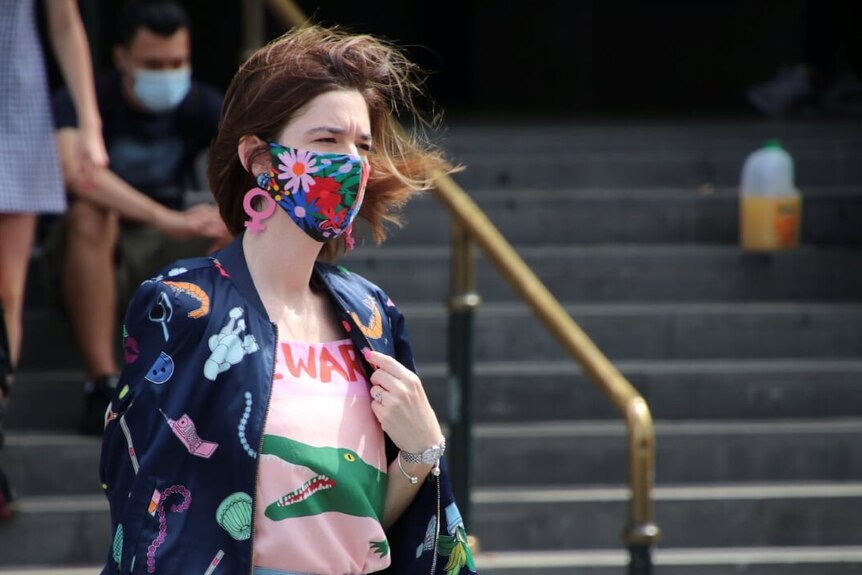 A woman wears a colourful mask, jacket, shirt and jeans as she walks past Flinders Street Station.