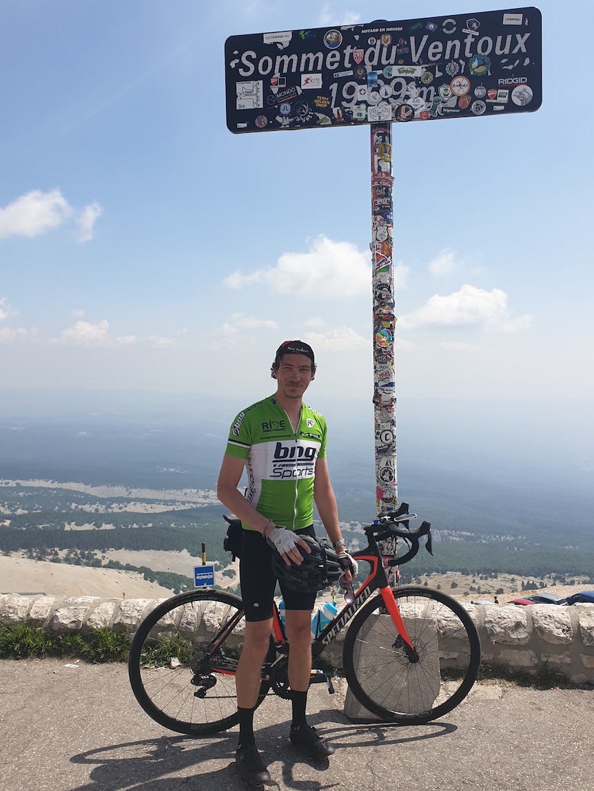 A man in green bicycle gear stands with a bike in front of a sign that reads "Somme du Ventoux".