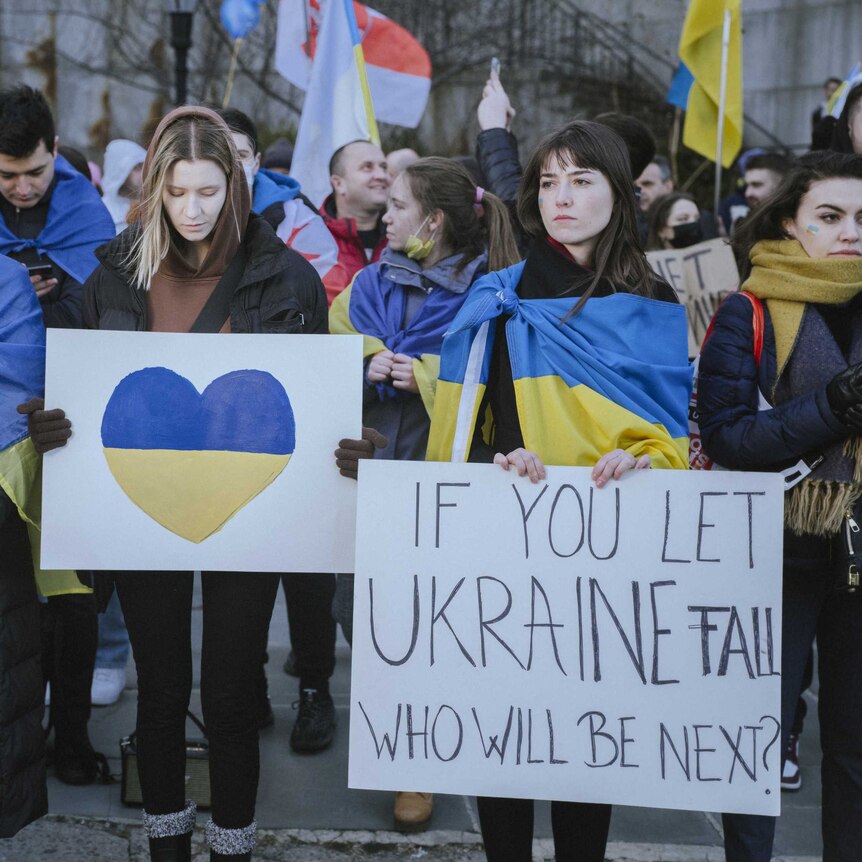protestor crowd, some wear Ukrainian flag. One has a sign with a heart, another says "if you let Ukraine fail who will be next"