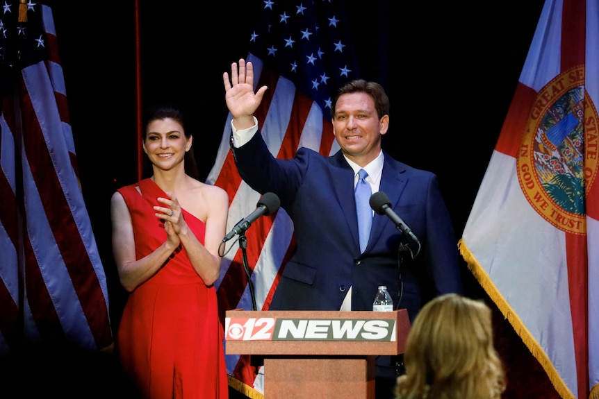 Ron Desantis, in a blue suit, waves at a crowd while his wife claps in a red dress