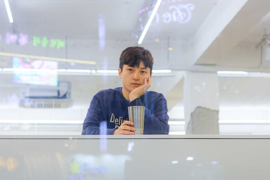 A man holding a milk shake cup sits behind a bar and gazes into the camera