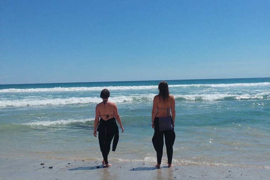 Two young women standing looking out to sea, wearing wetsuits with their backs to the camera