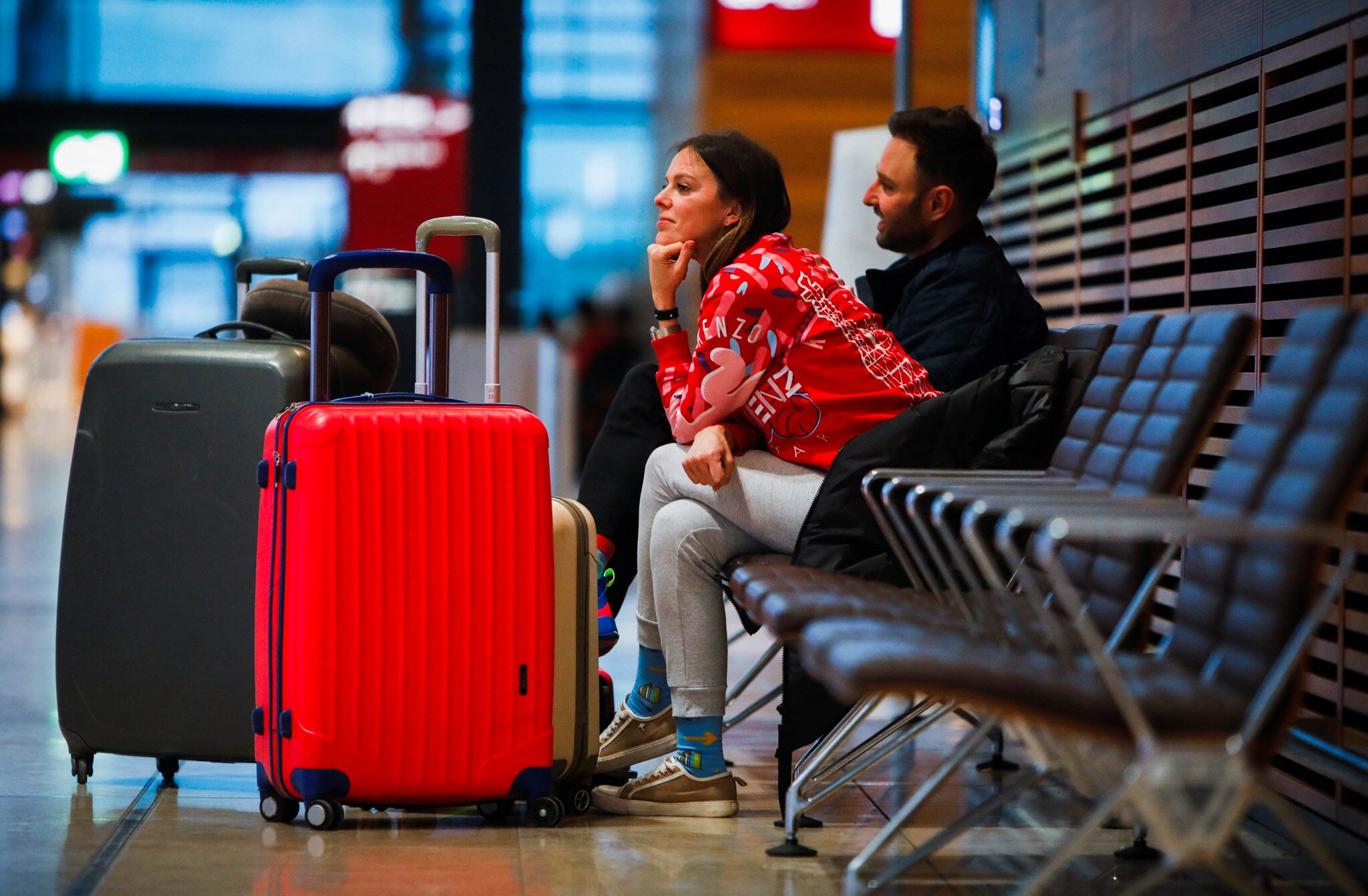 People wait with their luggage while sitting on bench seats at the airport.