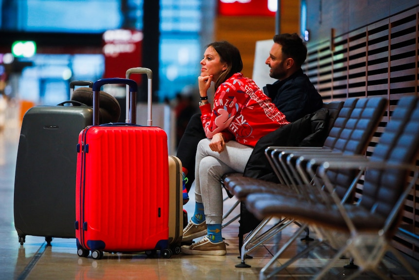 People wait with their luggage while sitting on bench seats at the airport.