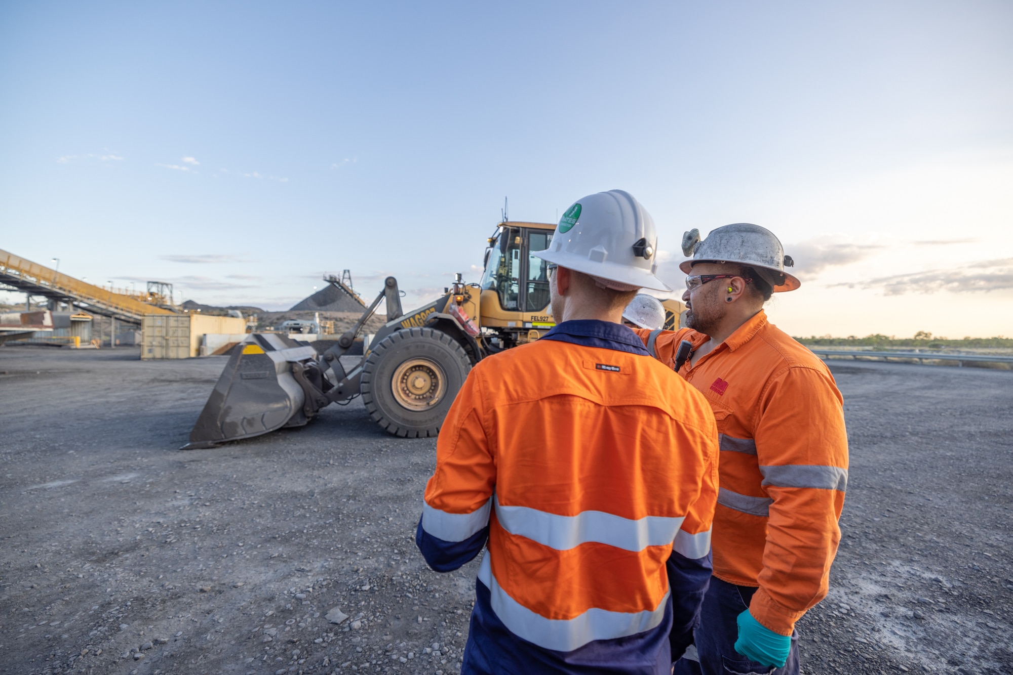 Two men standing on a mine site next to a grader and conveyer belt