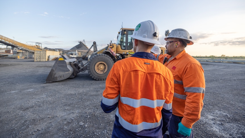 Two men standing on a mine site next to a grader and conveyer belt