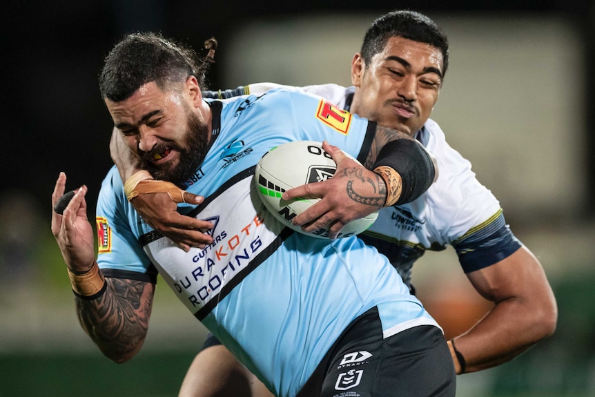 A Cronulla Sharks NRL players holds the ball with his left hand as he is tackled by a North Queensland opponent.