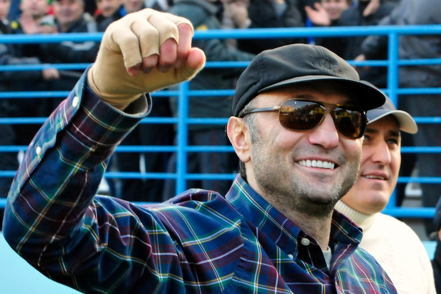 A man sunglasses and a hat and checked shirt raises his fist and smiles.
