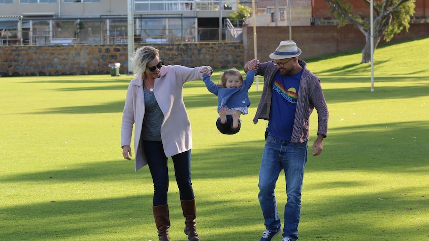 A man and a woman lift their smiling toddler up in the air while standing on a grass field