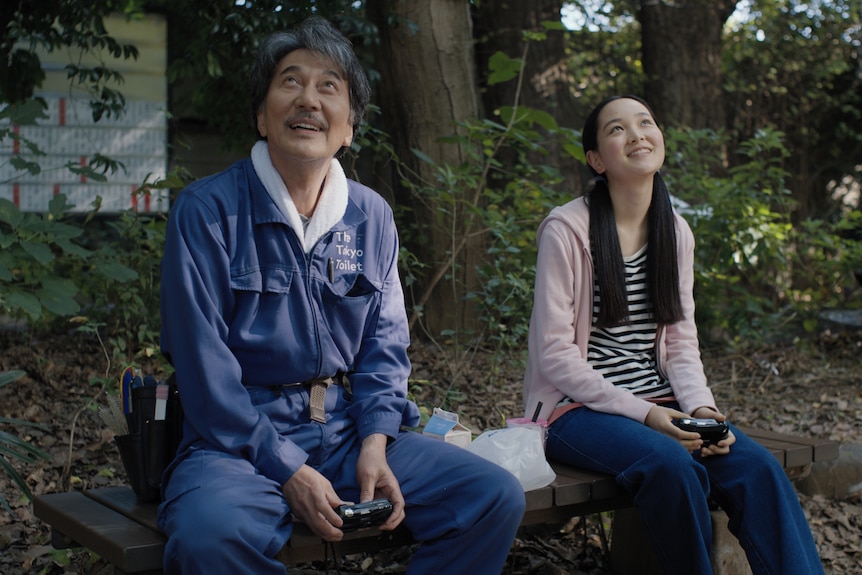 A film still of Kōji Yakusho and Arisa Nakano, two Japanese people, sitting together on a park bench, smiling as they look up.
