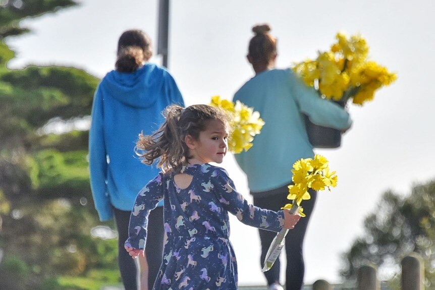 A young girl walks behind two women. All of them carrying bunches of daffodils as they walk up a street.