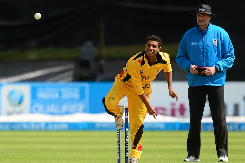 A spin bowler clad in yellow watches the flight of the ball after bowling in a cricket match as the umpire stands behind him.