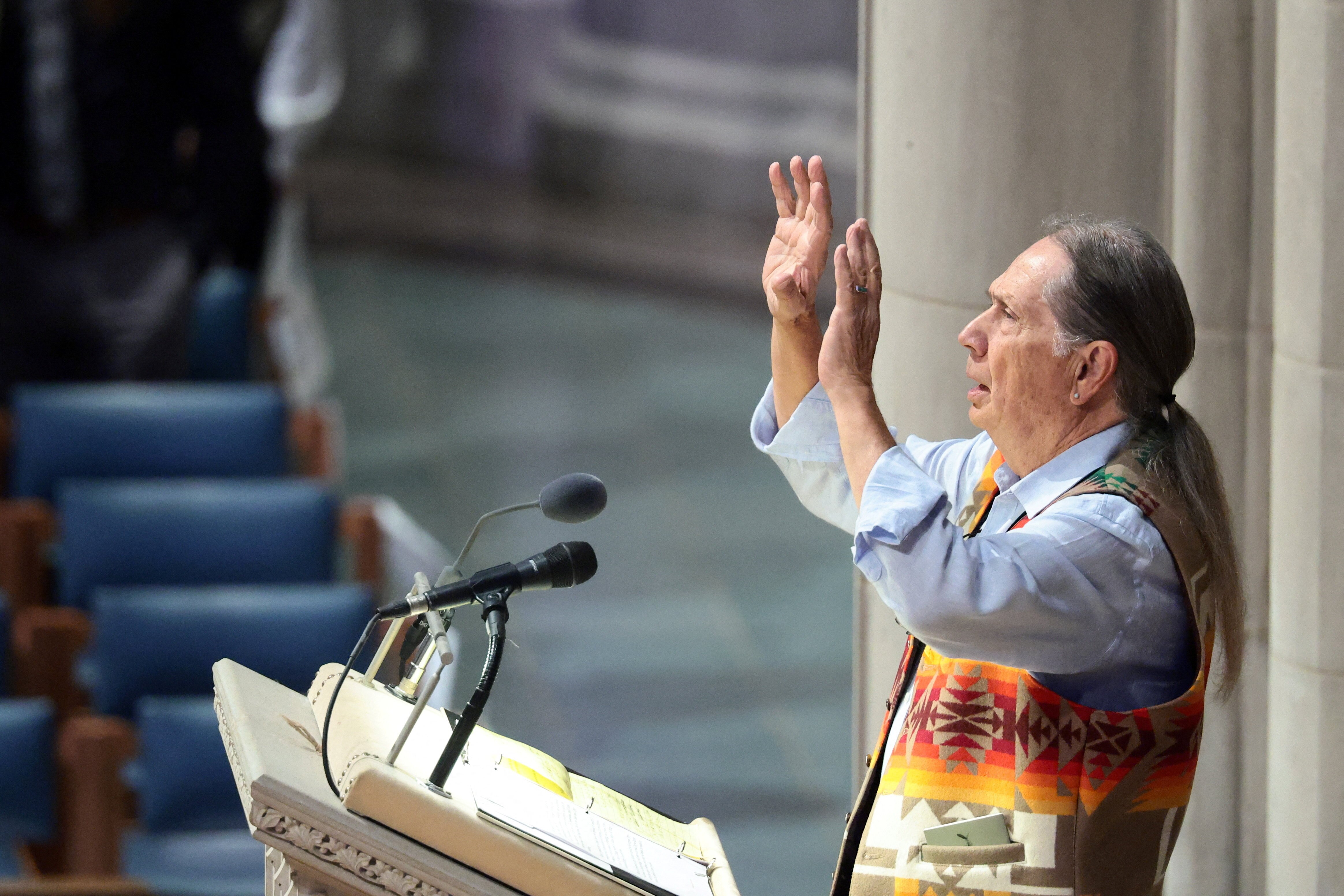 A man in a colourful vest holds up his hands while speaking a lectern in a cathedral.