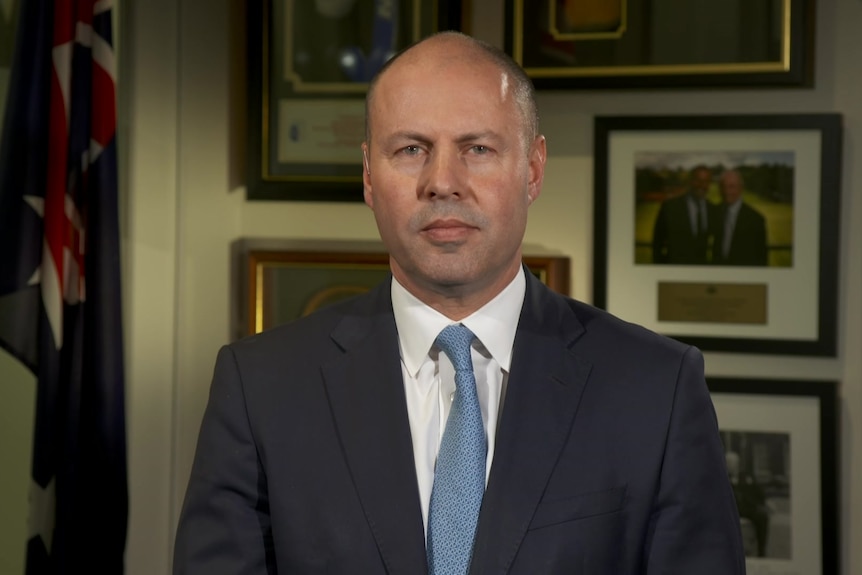 A man in a suit and blue tie stares into the camera. Behind him are portraits and the Australian flag.