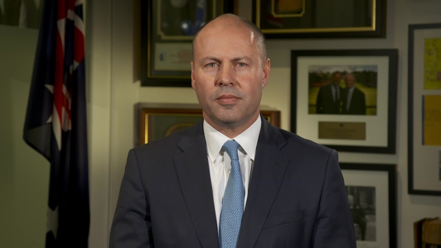 A man in a suit and blue tie stares into the camera. Behind him are portraits and the Australian flag.