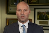 A man in a suit and blue tie stares into the camera. Behind him are portraits and the Australian flag.