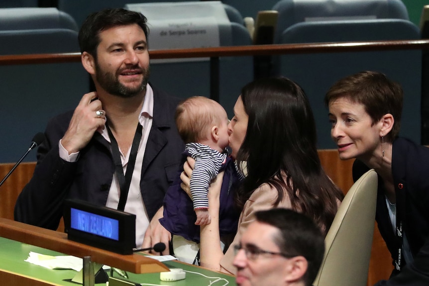 Jacinda Ardern kisses her baby's cheek while holding her up next to Clarke Gayford at the UN