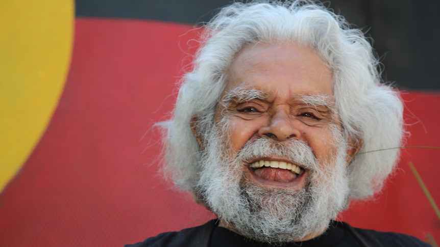 Uncle Jack Charles laughs, standing in front of an Aboriginal flag-coloured backdrop.
