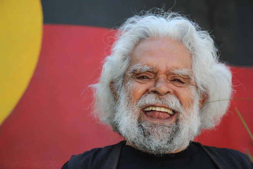 Uncle Jack Charles laughs, standing in front of an Aboriginal flag-coloured backdrop.