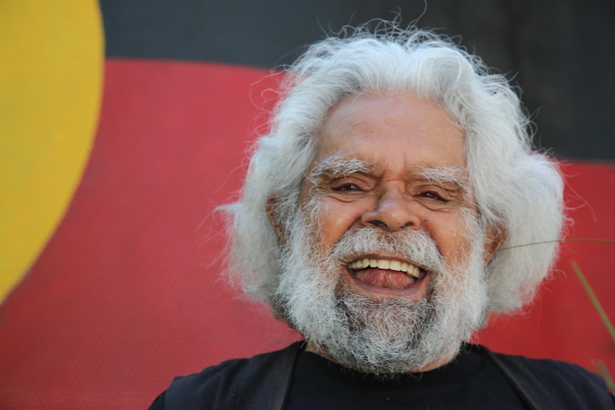 L'oncle Jack Charles rit, debout devant une toile de fond aux couleurs du drapeau autochtone.