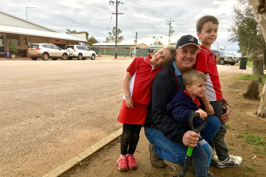 Senior man sits with a shovel along the edge of a  main street of country town with his young grandchildren.