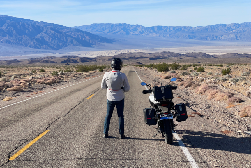 A man stands in the desert on a road with a motorcycle nearby