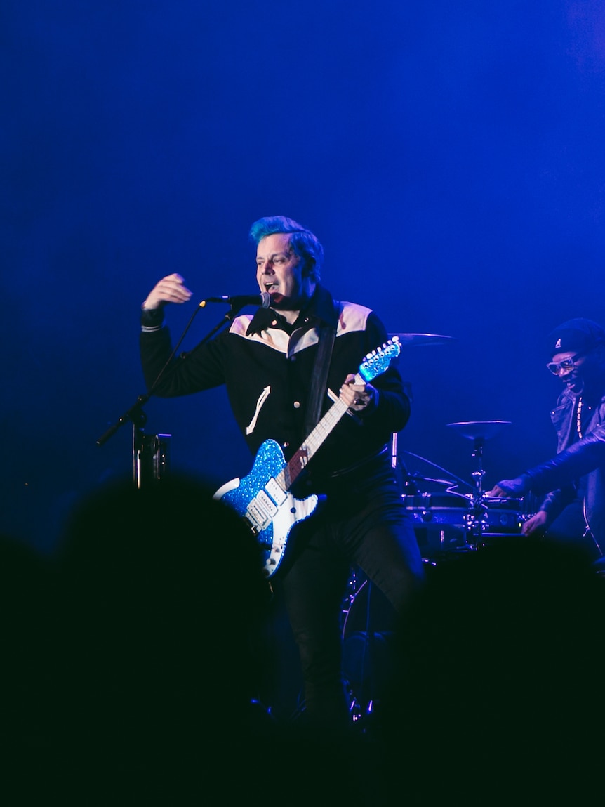Jack White sings into a mic, gestures to the crowd and holds a blue guitar