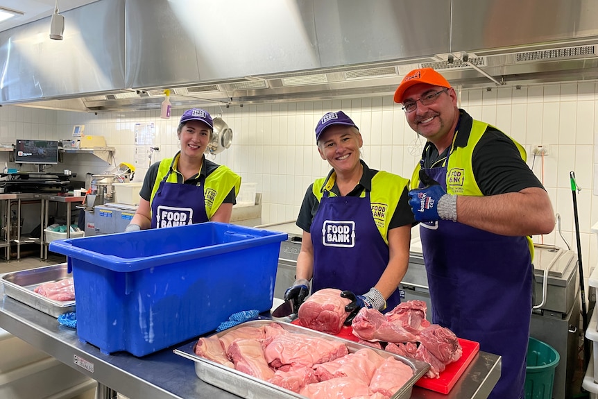Three Foodbank volunteers are in a kitchen smiling to the camera in the process of butchering meat. 