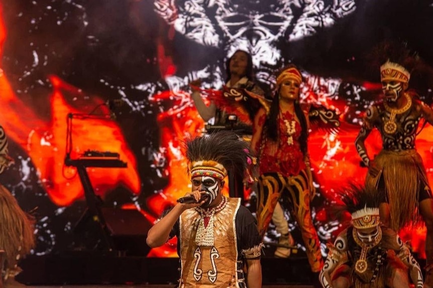 Dark skinned man in traditional make-up, dress and head covering on stage.