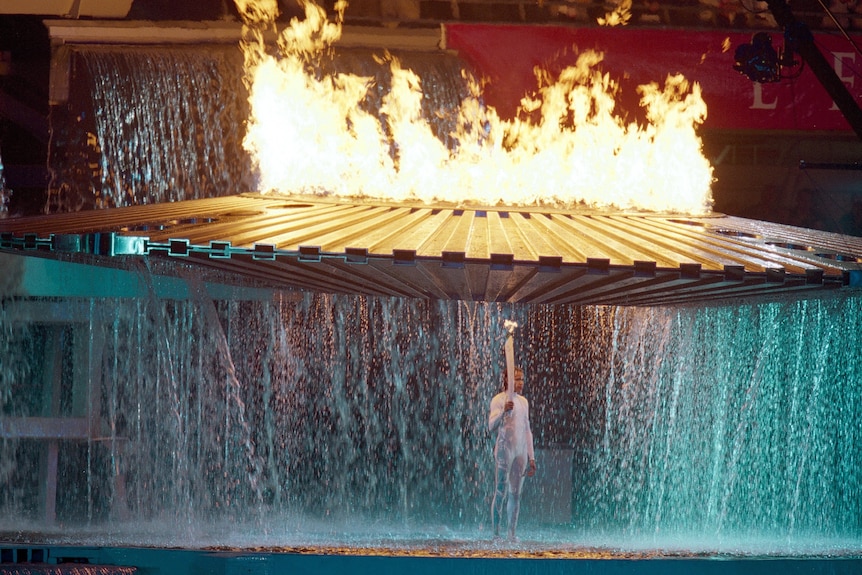 A woman in a white jumpsuit holds a flaming torch as water falls from a large cauldron above her head