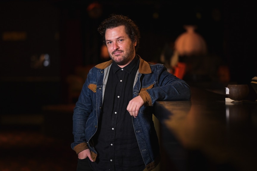 A man stands leaning on a bar with a dark background.