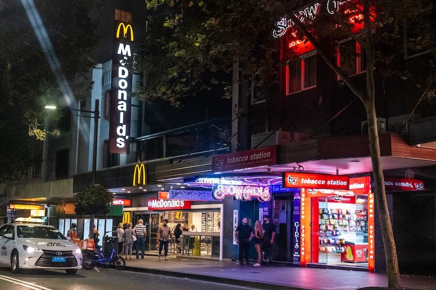 A group of people walk along an inner-city street which includes a McDonalds, a stripclub and a tobacconist