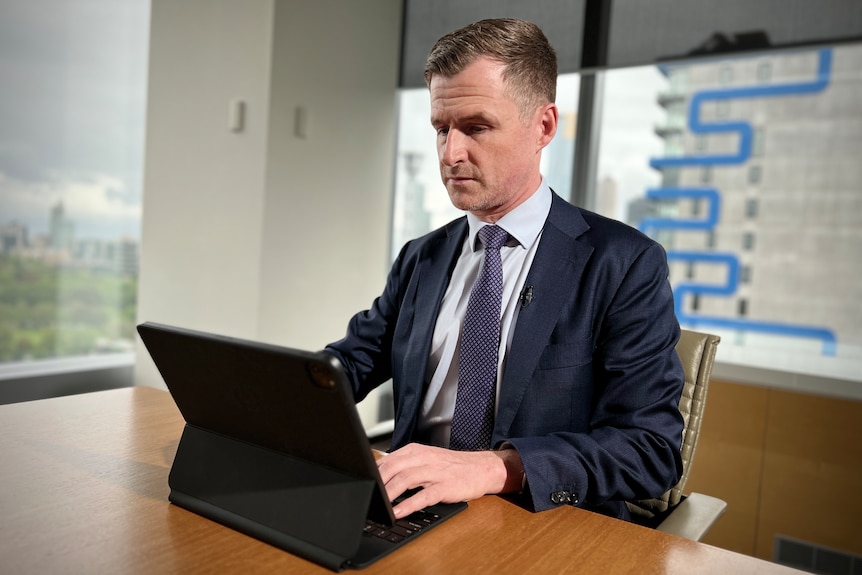 A man in a dark blue suit and tie sits at a table working on a laptop.