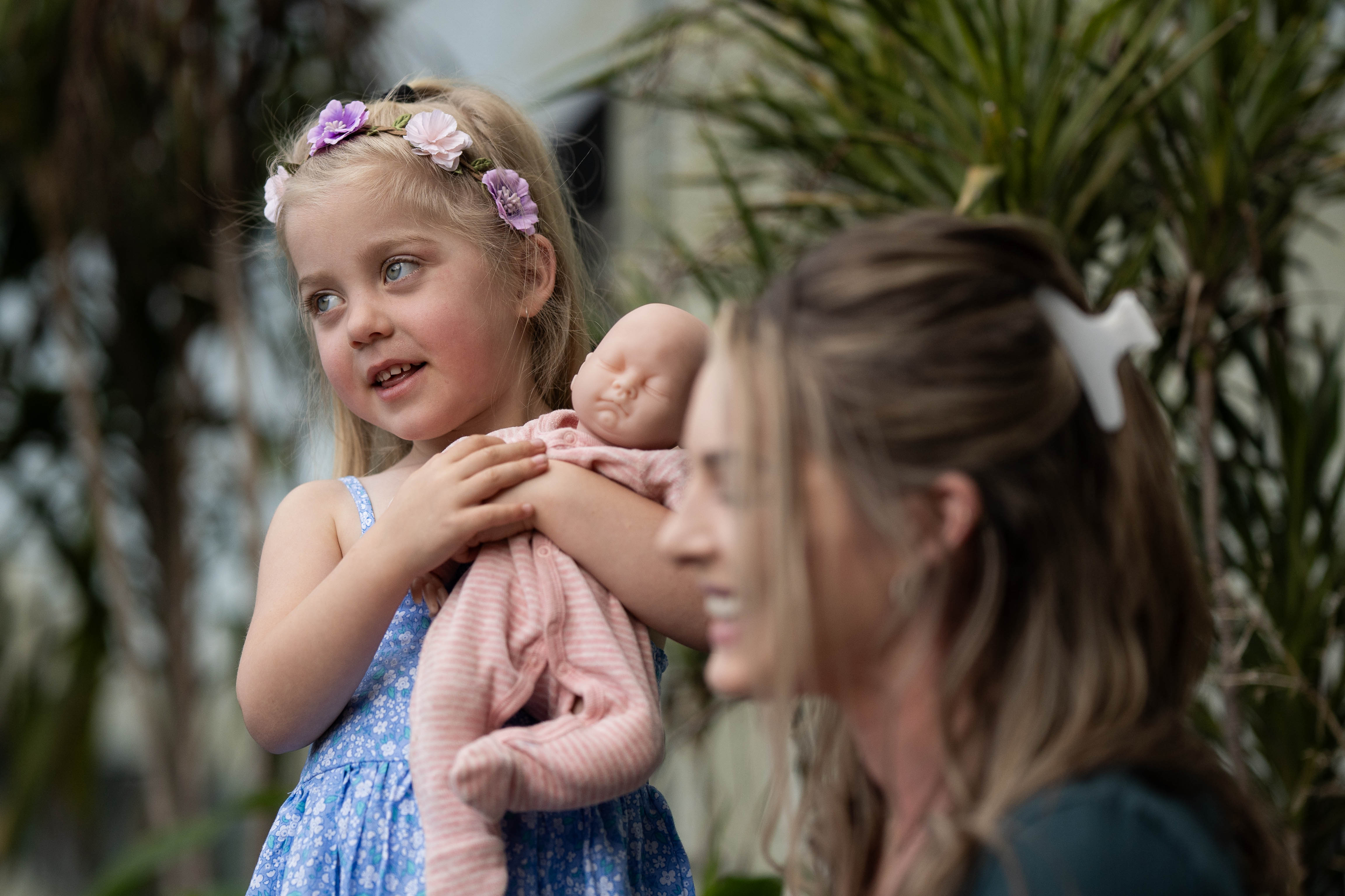 A young girl with flowers in her hair stands holding a doll next to her mother