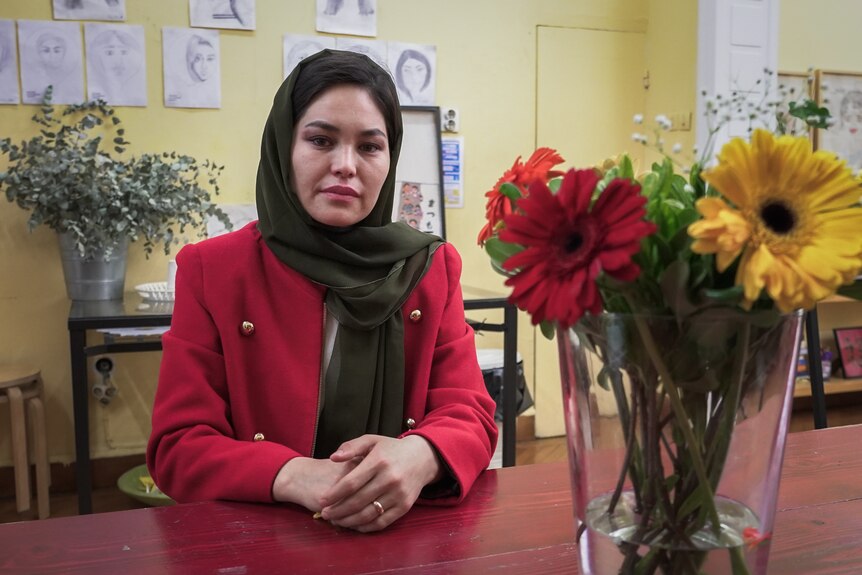A woman wearing a green hijab and pink jacket sits a desk with flowers on it.