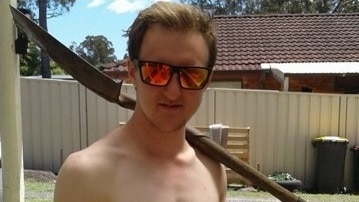 A shirtless young man wearing reflective sunglasses stands in a back yard with a shovel over his shoulder.