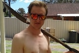 A shirtless young man wearing reflective sunglasses stands in a back yard with a shovel over his shoulder.