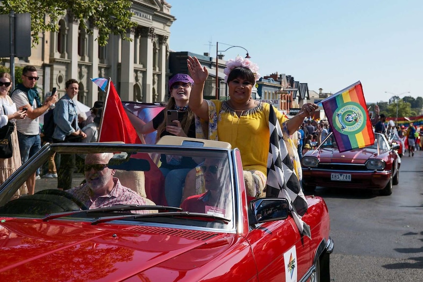 two women ride in a car during a parade and wave rainbow flags