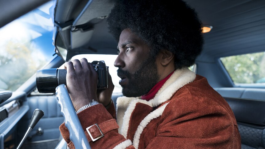 Colour close-up image of John David Washington holding binoculars and sitting in a car.