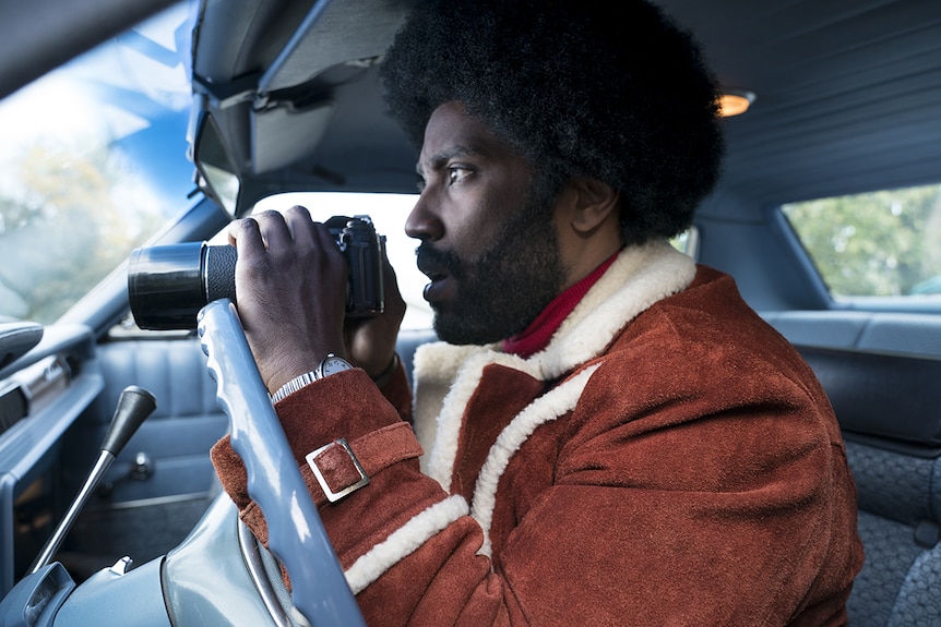 Colour close-up image of John David Washington holding binoculars and sitting in a car.