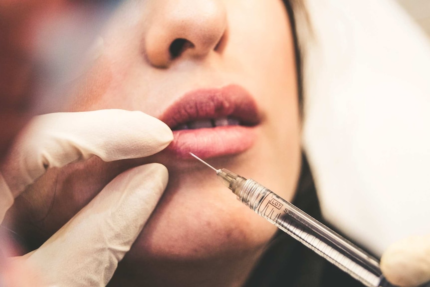 A surgeon injects filler into a young woman's lips