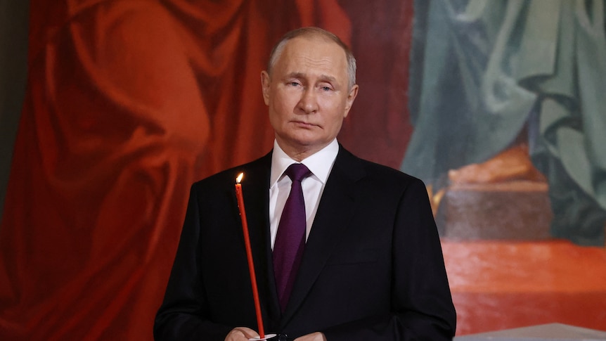 Vladimir Putin, a balding man dressed in a dark suit, poses for a photo in a church holding a long red candle