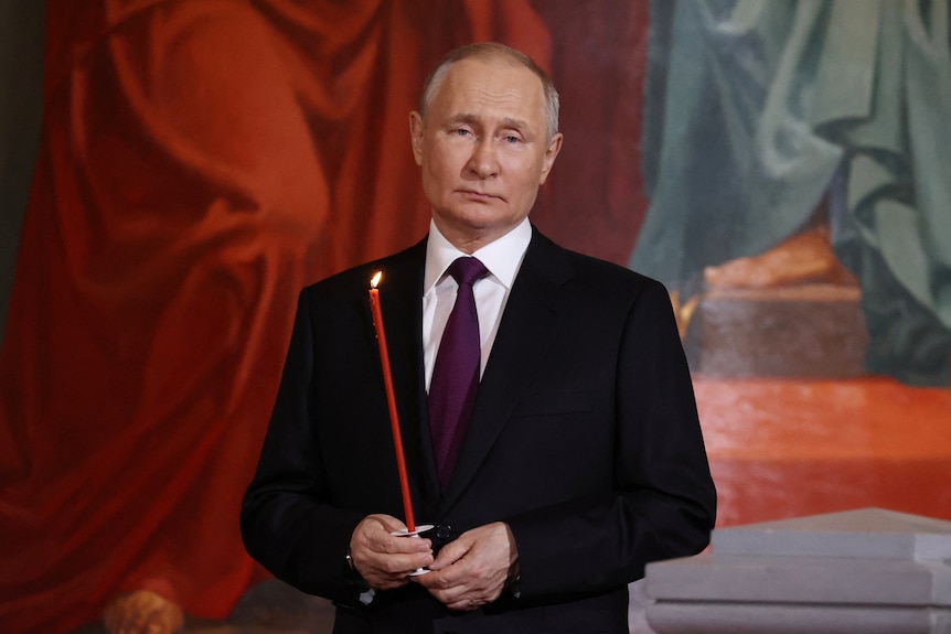 Vladimir Putin, a balding man dressed in a dark suit, poses for a photo in a church holding a long red candle