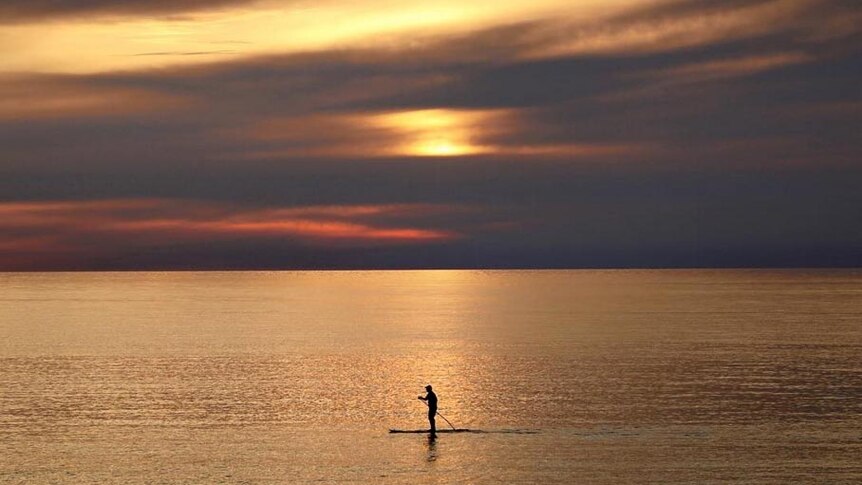 Elevated view of a stand up paddler making their way across the ocean as the sun sets on the horizon.