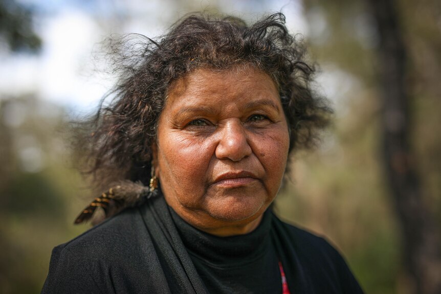A solemn Aboriginal woman looks directly at the camera, wears black, and a feather in her curly hair.