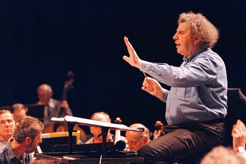 A man with frizzy hair gestures while conducting an orchestra