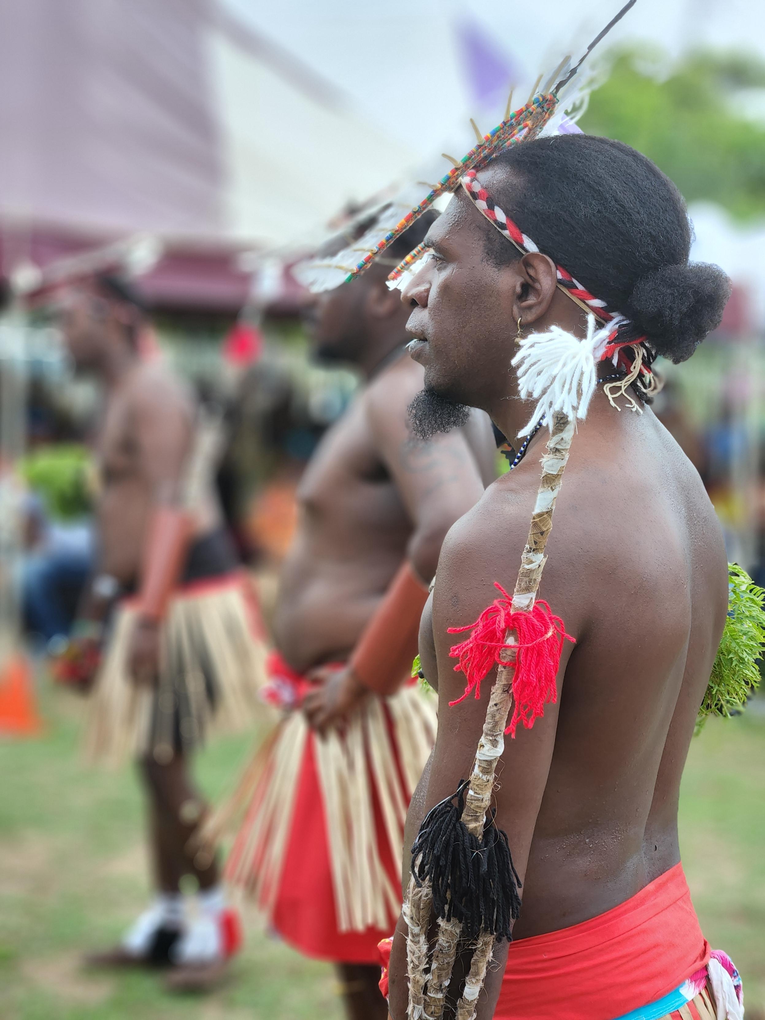 A First Nations man stands wearing woven straw headgear and holding long, thin stick, staring ahead with slight smile.