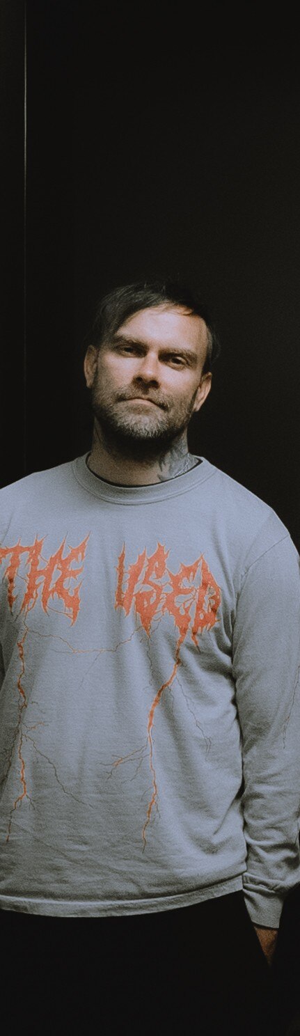 Bert McCracken from emo-punk band The Used wears a grey band jumper with 'The Used' written in orange writing on the front