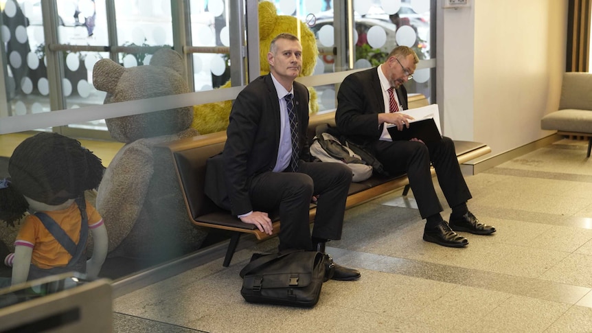 Two men in suits wait on a bench seat.